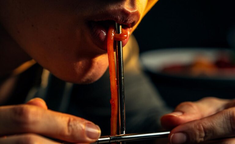 Illustration von einer Person, die mit konzentriertem Gesicht eine einzelne Spaghetti durch einen metallenen Strohhalm ansaugt, vor einem minimalistischen, feinen Tischsetting.